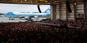 Eine Menschenmenge steht in einer Halle, die auf einer Seite geöffnet ist, dort steht ein weiß-blaues Flugzeug