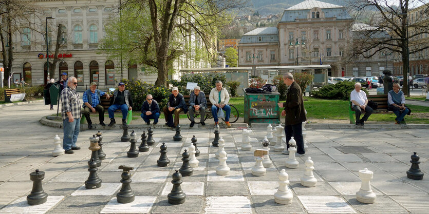 Männer sitzen auf einer Bank auf einem Platz. Vor ihnen ist ein großes Spiel mit Figuren aufgebaut