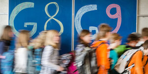 Kinder laufen durch einen Klassenraum. Im Hintergrund ist eine Tafel zu sehen, auf der "G8" und "G9" steht