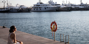 Eine Frau mit Kopfhörern im Ohr sitzt an einem Yachthafen in der Sonne