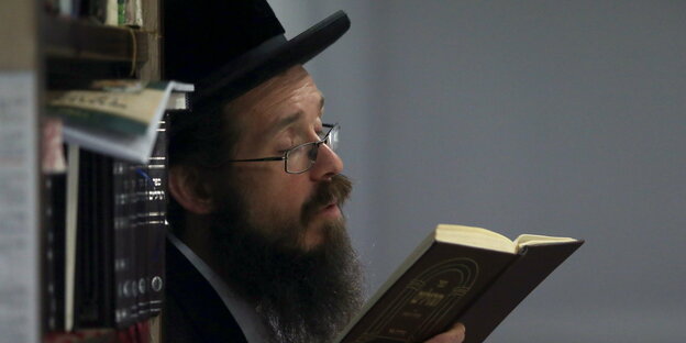 Ein orthodoxer Jude mit Hut, Brille und langem Bart, lehtn an einem Bücherregal, hält ein Buch und betet