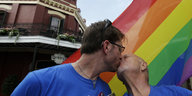 Zwei Männer, beide in blauen T-Shorts, küssen sich vor einer Regenbogen-Flagge