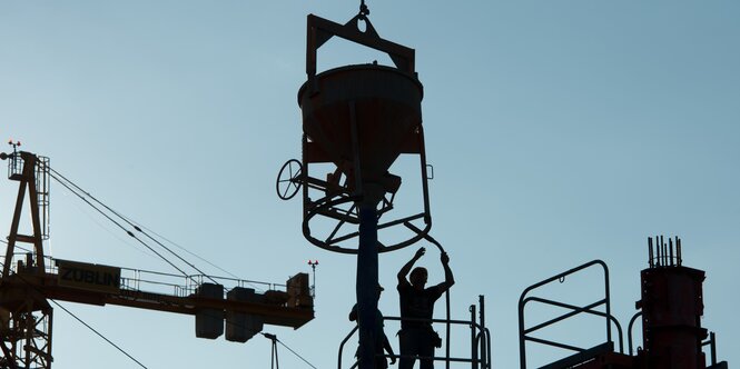 Vor Stahlblauem Himmel zeichnen sich schwarz Kräne und Zementbehälter in luftiger Höhe aus