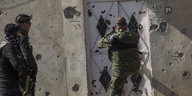 schwer bewaffnet stehen drei Soldaten vor einer von Schüssen durchlöcherten Hauswand