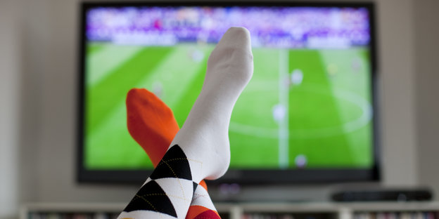 Überschlagene Beine und im Hintergrund ein Fernsehen, auf dem ein Fußballspiel gezeigt wird