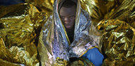 Ein Mensch hüllt sich in eine gold glitzernde Rettungsdecke, umgeben von weiteren Rettungsdecken