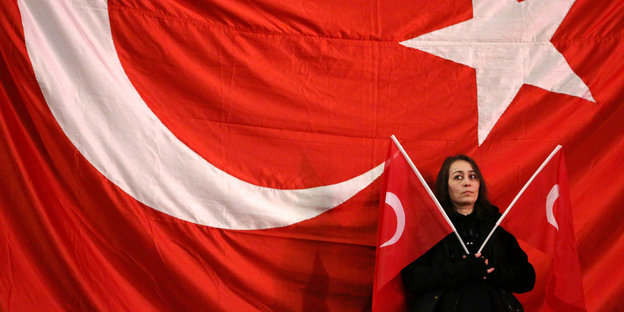 Eine Frau steht vor einer großen Türkeiflagge und hält zwei kleinere Flaggen in den Händen