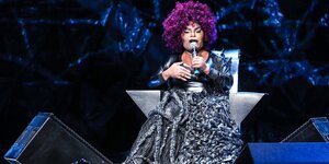 Eine Frau mit einer lilafarbenen Afro-Frisur sitzt auf einer Bühne und ein schwarzes Kleid mit vielen Verflechtungen an