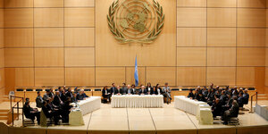 Menschen sitzen in einem großen Saal um Tische unter einem riesigen UN-Logo