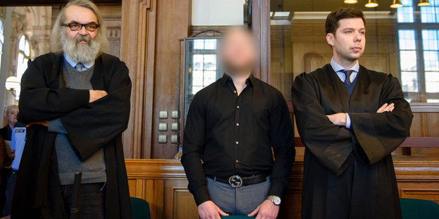 drei Männer in einem Gerichtssaal, das Gesicht des mittleren Mannes ist verpixelt