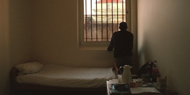 Mann steht an einem vergitterten Fenster in einem karg eingerichteten Raum
