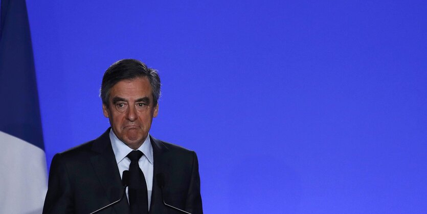 Francois Fillon steht vor einer blauen Wand und zieht die Mundwinkel nach unten