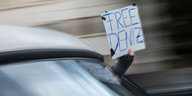 Verwischtes Bild, dass Geschwindigkeit zeigt. Jemand hält ein Schild aus dem Fenster. Darauf steht: Free Deniz