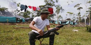 Ein Mann sitzt im Freien auf einem Hocker und macht etwas mit seinem Gewehr