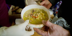 Jemand überreicht einer anderen Person einen Plastikteller voll Suppe