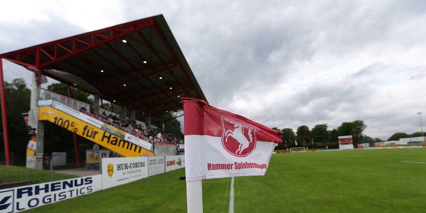 Vor einer Zuschauertribüne steht eine rot-weiße Fahne auf einem grünen Fußballfeld