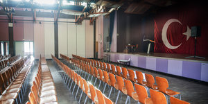 In einer Halle stehen Reihen orangefarbener Stühle, auf einer Bühne hängt eine Fahne der Türkei