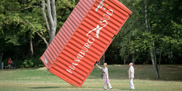 Ein roter Container steht auf einer Spitze auf einer Wiese, daneben zwei Menschen