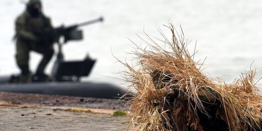 Ein Soldat in Strohtarnung liegt auf dem Boden, dahinter sitzt einer mit einem Gewehr