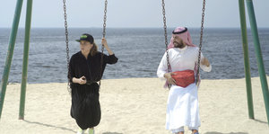 Eine Frau im langen schwarzen Kleid und Käppi und ein Mann in traditionell saudischer Kleidung sitzen am Strand nebeneinander auf Schaukeln