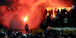 eine Massenschlägerei bei Nacht in einem Fußballsadion, im Vordergrund Polizisten, dazwischen ein Feuer und roter Qualm