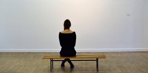 Eine Museumsbesucherin sitzt auf einer schmalen Bank und blickt auf eine weiße Wand