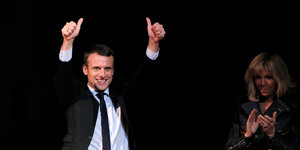 Emmanuel Macron steht vor schwarzem Hintergrund und hat beide Arme, inklusive die Daumen nach oben gestreckt