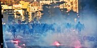Im Hintergrund die Fassaden mehrerer Häuser in Neapel, im Vordergrund eine Menschenmenge vor denen es raucht und kleine Flammen brennen