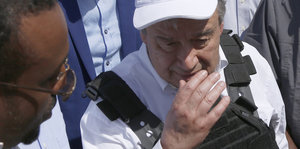 Ein älterer Mann in weißem T-Shirt, schwarzer Weste und weißer Basecap hält sich die Hand vors Gesicht