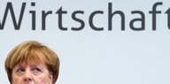 "Wirtschaft" steht an einer hellen Wand, darunter ist Merkels Gesich halb zu sehen