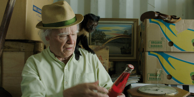 Ein alter Mann guckt eine Flasche rote Brause an. Ihm sitzt ein Äffchen auf der Schulter und guckt auch auf die Brauseflasche.
