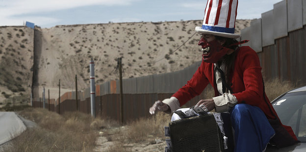 Mann als Onkel Sam verkleidet - mit einem Koffer voller Dollar in der Wüste, durch die sich ein Grenzzaun zieht