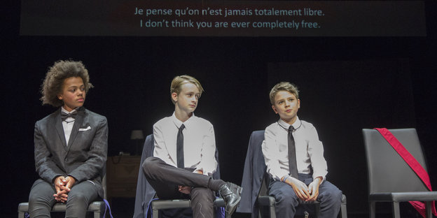 Drei Jungen sitzen wie Erwachsene gekleidet auf der Bühne