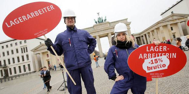 Ein Mann und eine Frau in Arbeitskleidung mit Plakaten vor dem Brandenburger Tor