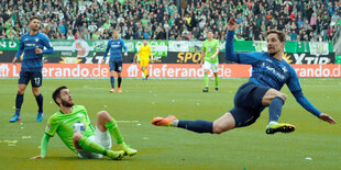 Zwei Fußballspieler beim Spiel, einer sitzt auf dem Rasen, der andere befindet sich gerade in einer Flugphase