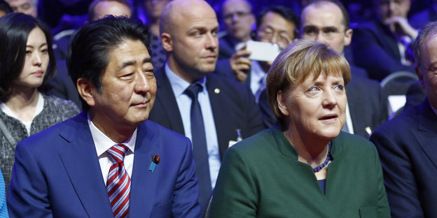 Shinzo Abe, Angela Merkel und diverse Anzugträger sitzen in mehreren Stuhlreihen hintereinander