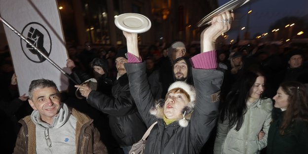 Eine Frau schlägt in einer Menge von Demonstranten zwei Topfdeckel gegeneinander