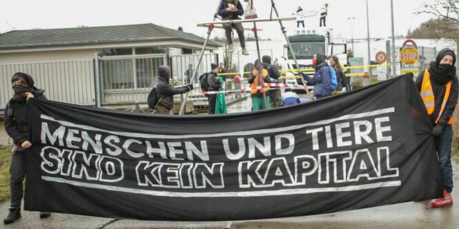 Die Demonstranten blockieren die Zufahrt des Schlachthofes und spannen ein Tuch mit der Aufschrift "Menschen und Tiere sind kein Kapital".