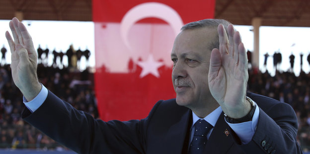 Erdogan steht vor einer türkischen Flagge und hebt messianisch beide Hände