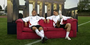 Die Fußballer Podolski und Schweinsteiger sitzen nebeneinander auf einem Sofa auf einem Fußballrasen vor einer Fotokulisse
