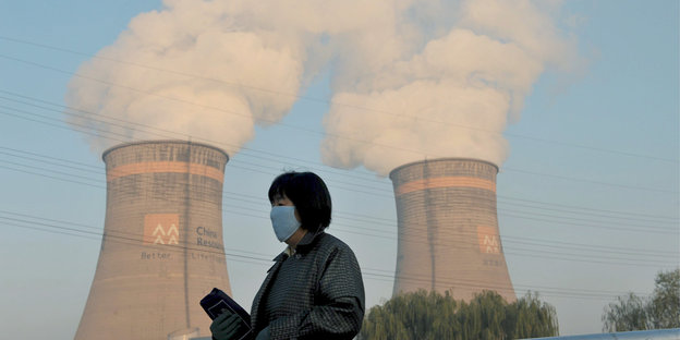 Eine Frau mit Atemschutzmaske vor Türmen eines Kohlekraftwerks