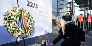 Eine Frau, die in schwarz gekleidet ist, legt einen Blumenstrauß vor einem einem großen Blumenkranz nieder