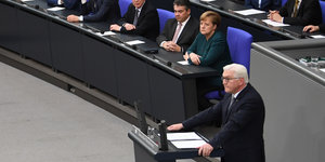 Ein Mann mit weißen Haaren steht am Rednerpult, weitere Menschen sitzen im Hintergrund und hören ihm zu