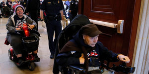 Zwei Personen in Rollstühlen werden von einem Polizisten durch eine Tür begleitet