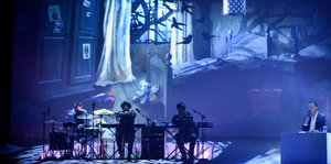 Auf einer Bühne spielt eine kleine Gruppe Musiker ein Stück, rechts im Bild sitzt ein Mann vor einem Pult