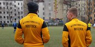 Zwei Männer stehen mit dem Rücken zur Kamera und blicken auf ein Fußballspiel. Auf ihren Jacken steht „Internationaler FC Rostock“