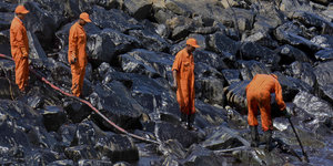 Vier Männer mit orangefarbener Schutzkleidung reinigen einen Steinstrand, der mit Erdöl verunreinigt ist.