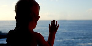 Ein Baby steht an einem Fenster, dahinter das Meer