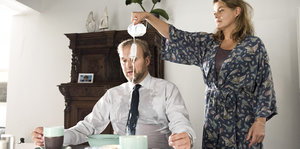 Eine Frau gießt stehend einem vor ihr sitzenden Mann ein Kännchen Milch über den Kopf