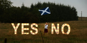 Ein Mann im Schottenrock schwenkt eine schottsche Fahne. Er steht auf einer Wiese zwischen zwei großen Leuchtröhren, die die Buchstaben „Yes“ und „No“ abbilden, dahinter Wald